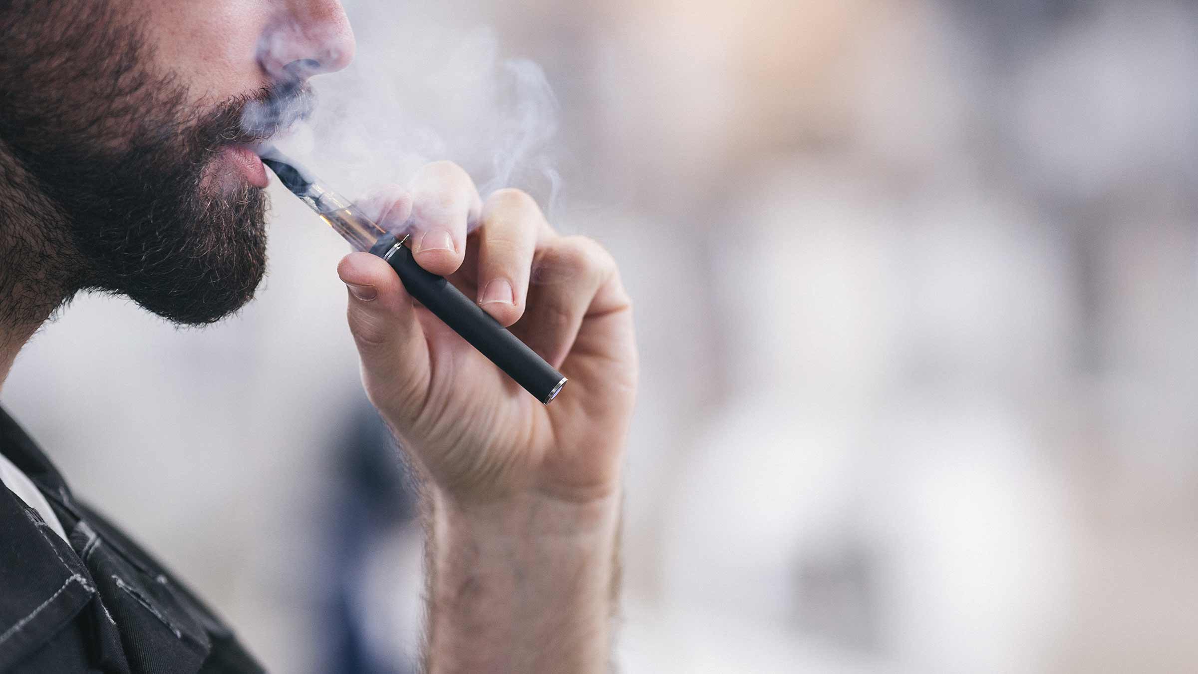 A bearded man vaping outdoors.